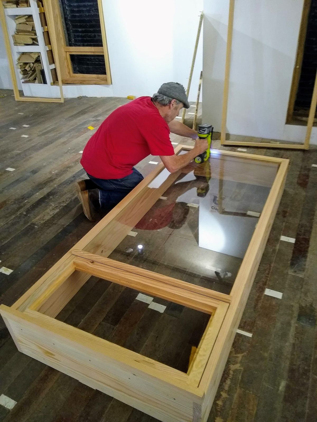 Schrijnwerker in rood shirt knielt bij een houten raamkader met glas op de houten vloer van een werkruimte